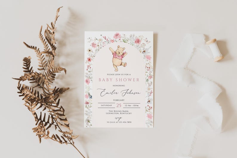 Pode incluir: Um convite para ch&aacute; de beb&ecirc; com Ursinho Pooh, rodeado por um arco floral. O convite inclui o texto "Please join us for a Baby Shower honoring Emilia Johnson" e detalhes do evento. Um ramo de samambaia seca e fita branca tamb&eacute;m est&atilde;o na imagem.