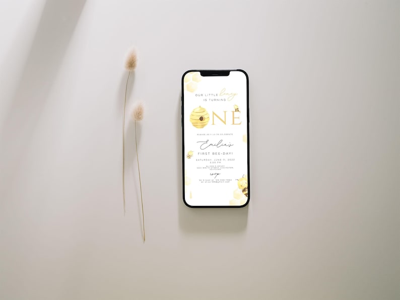 Może przedstawiać: Smartfon wyświetlający zaproszenie na pierwsze urodziny z motywem pszcz&oacute;ł. Zaproszenie przedstawia plaster miodu z pszczołą i cyfrą "jeden" w złotych literach. Tekst brzmi "Our little bee is turning one" i "Emilias First Bee-Day".