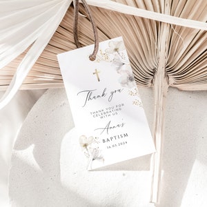 Puede incluir: Una etiqueta de bautizo blanca con una cruz dorada y detalles florales. La etiqueta dice "Thank you" e incluye el nombre "Anna's" y la fecha "16.03.2024". La etiqueta est&aacute; suspendida por un cord&oacute;n marr&oacute;n sobre un fondo neutro.