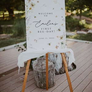 May include: A white sign with a floral design and bees. The sign says "Welcome to Emilia's First Bee-Day! June 11, 2022".