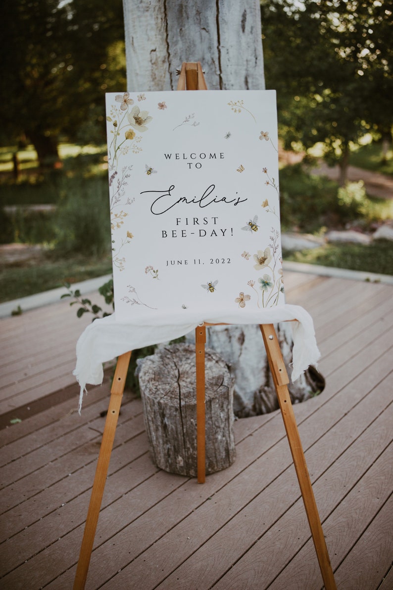 May include: A white sign with a floral design and bees. The sign says "Welcome to Emilia's First Bee-Day! June 11, 2022".