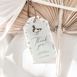 May include: A rectangular gift tag with a watercolor illustration of a duck, surrounded by floral elements. The tag reads "Thank you" and includes baby shower details. The tag is suspended by a brown cord.