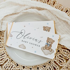 May include: White chocolate bar wrapper with a gold border. The wrapper features a blue and brown teddy bear sitting on a cloud with blue stars and the text "Olivia's Baby Shower".
