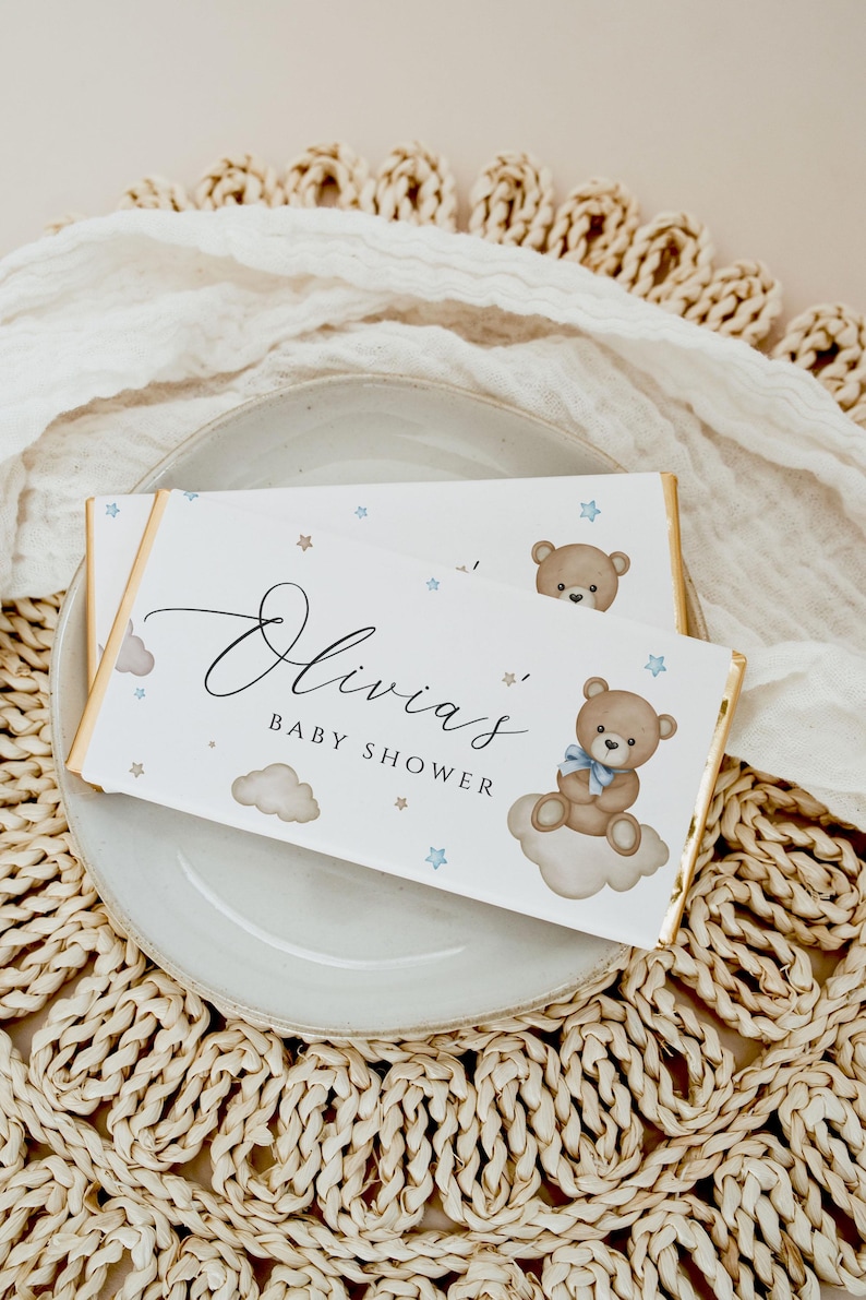 May include: White chocolate bar wrapper with a gold border. The wrapper features a blue and brown teddy bear sitting on a cloud with blue stars and the text "Olivia's Baby Shower".
