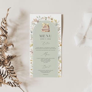 May include: A green and white menu card with a watercolor illustration of a stack of books and floral accents. The menu card has the text "MENU" and "JUNE 11, 2022" at the top. The menu includes sections for "Entrees", "Main", and "Dessert".