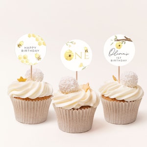 Op de afbeelding: Drie cupcakes versierd met witte frosting en bedekt met cupcake-toppers met een bijenmotief. De toppers hebben een honingraatontwerp, een bijenkorf met het nummer "EEN" en de tekst "Olivia's 1st Birthday".