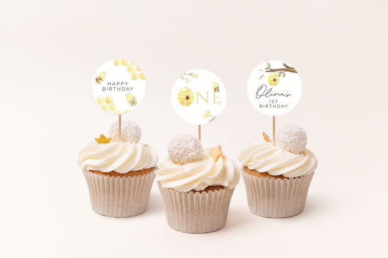 Op de afbeelding: Drie cupcakes versierd met witte frosting en bedekt met cupcake-toppers met een bijenmotief. De toppers hebben een honingraatontwerp, een bijenkorf met het nummer "EEN" en de tekst "Olivia's 1st Birthday".