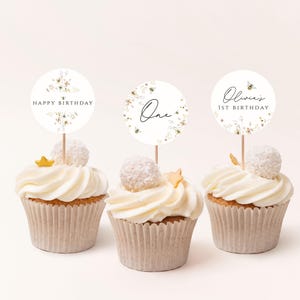 May include: Three white cupcakes with white frosting and gold sprinkles. Each cupcake has a round, white paper topper with floral designs and black text. The toppers read "Happy Birthday", "One", and "Olivia's 1st Birthday".