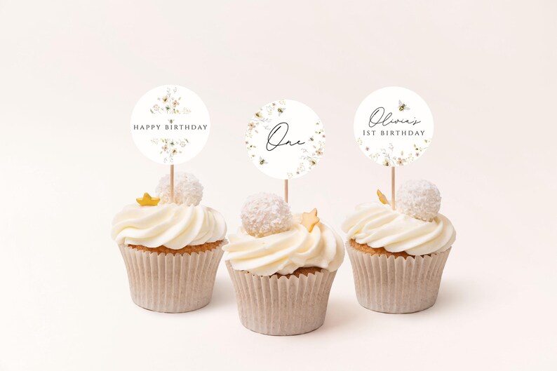 May include: Three white cupcakes with white frosting and gold sprinkles. Each cupcake has a round, white paper topper with floral designs and black text. The toppers read "Happy Birthday", "One", and "Olivia's 1st Birthday".