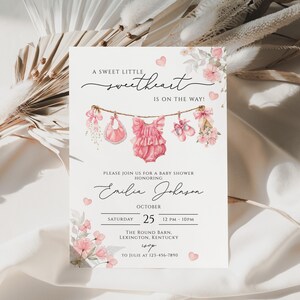 Może przedstawiać: Zaproszenie na baby shower z akwarelowymi ilustracjami ubranek dla dzieci i kwiatowymi akcentami. Zaproszenie zawiera tekst "A Sweet Little Sweetheart Is On The Way!" oraz szczegóły wydarzenia, w tym imię, datę, godzinę i miejsce.