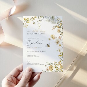 K&ouml;nnte beinhalten: Eine wei&szlig;e Einladungskarte mit einem floralen Design mit gelben und gr&uuml;nen Blumen, Bienen und Bl&auml;ttern. Der Text auf der Karte lautet "Unser kleiner Honig wird ein Jahr alt!" und "Emilias erster Bienentag!" Das Datum der Veranstaltung ist Samstag, der 18. Juni 2022.
