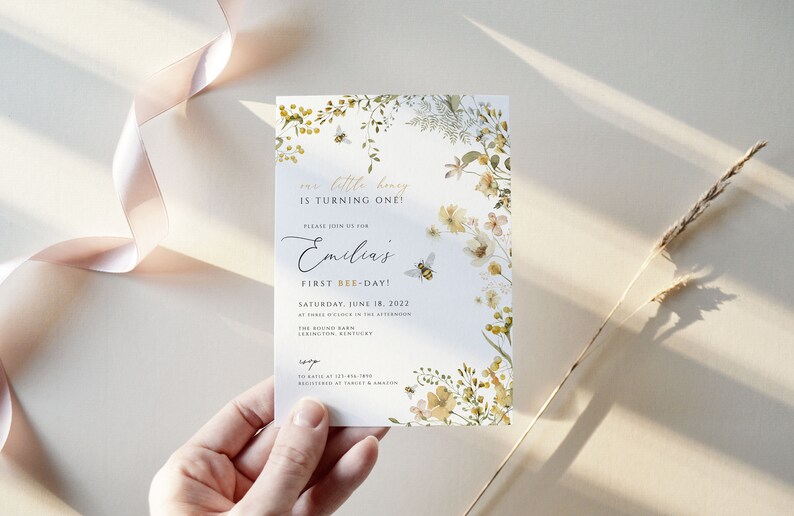 K&ouml;nnte beinhalten: Eine wei&szlig;e Einladungskarte mit einem floralen Design mit gelben und gr&uuml;nen Blumen, Bienen und Bl&auml;ttern. Der Text auf der Karte lautet "Unser kleiner Honig wird ein Jahr alt!" und "Emilias erster Bienentag!" Das Datum der Veranstaltung ist Samstag, der 18. Juni 2022.