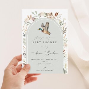 May include: A light blue baby shower invitation with a watercolor illustration of a duck flying through an archway of greenery. The text reads "Please join us for a Baby Shower honoring Anna Brooks, Saturday, December 19, 2022, 1:00 PM, The Round Barn, Lexington, Kentucky."