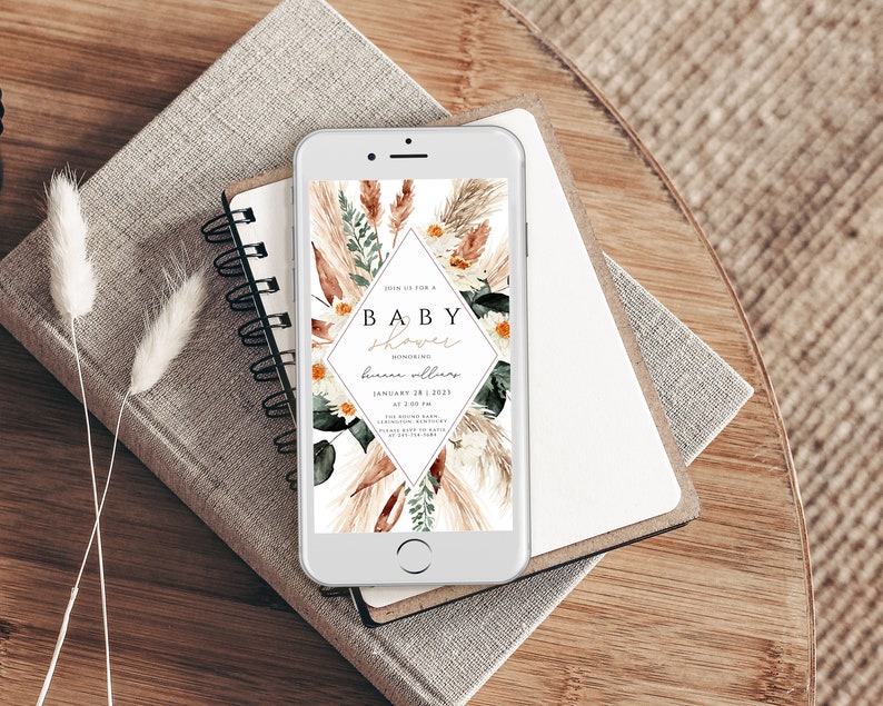 May include: A digital invitation on a smartphone screen for a baby shower. The invitation features a floral design with pampas grass, eucalyptus leaves, and white daisies. The text on the invitation reads "Join us for a Baby Shower Honoring [name] January 28, 2023 at 2:00 PM [address]"