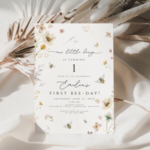 May include: A white invitation card with a floral design and the text "one little bug is turning 1. Join us to celebrate Emilia's First Bee-Day! Saturday, June 11, 2022 1:00 PM Allison's House 1234 White Street, Lexington, Kentucky RSVP to Allison at 123-456-7890"