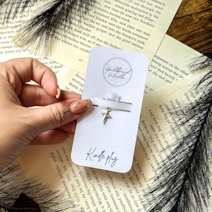 Op de afbeelding: Een zilveren Kindle-stekker met een sterbedel, gepresenteerd op een witte kaart met de tekst "Kindle plug". De kaart bevat ook een cirkelvormig logo met de woorden "Spellbound Scrolls". De achtergrond bevat open boeken.
