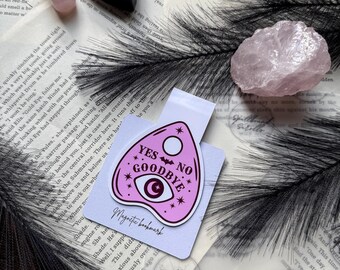 May include: A pink Ouija board-shaped magnetic bookmark with the words "YES NO GOODBYE" and an eye and crescent moon design. The bookmark is on a white card with the words "Magnetic bookmark" printed on it. Other items include a rose quartz crystal and a black feather.