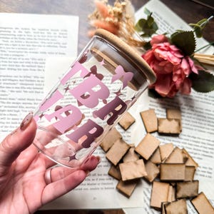 Könnte beinhalten: Ein klares Glas mit Holzdeckel, auf dem das Wort "LIBRARY" in rosa Buchstaben und Stern- und Schleifenakzenten steht. Das Glas wird in einer Hand gehalten, mit einem Hintergrund aus einer Buchseite und dekorativen Blumen.