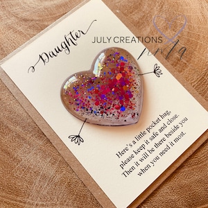 May include: A white card with the text "Daughter" and "JULY CREATIONS" in black cursive writing. A clear heart-shaped resin ornament filled with colorful glitter is attached to the card with a black arrow. The card reads "Here's a little pocket hug, please keep it safe and close. Then it will be there beside you when you need it most."