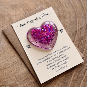 May include: A heart-shaped resin token filled with pink and purple glitter, attached to a cream-coloured card. The card features the text "One Day at a Time" and a poem about support, ending with "Love Mum xx," all resting on a brown envelope.