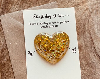 May include: A card with the text "First day at Uni" and "Here's a little hug to remind you how amazing you are." A gold glitter heart with a small arrow on each side is attached to the card.
