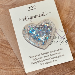 May include: A heart-shaped resin pin with iridescent glitter, set on a white card with the word "Alignment" and the number "222". The card also includes inspirational text. The pin is on a brown envelope.