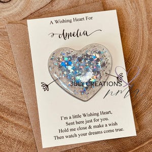 Könnte beinhalten: Ein herzförmiger Wunschstein mit silbernen und blauen Glitzer, auf einer Karte mit der Aufschrift "A Wishing Heart For Amelia" und "July Creations". Die Karte befindet sich auf einem braunen Umschlag mit einer Nachricht.