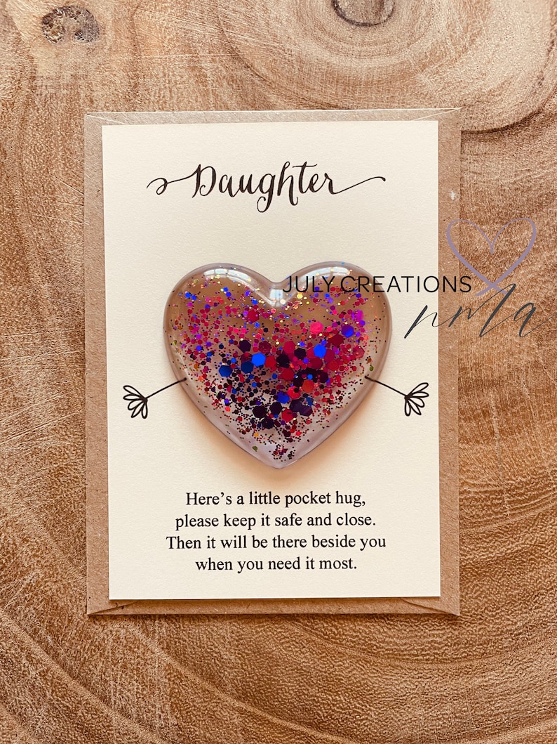 May include: A card with the word "Daughter" written in cursive on it. A small heart-shaped resin ornament with pink, blue, and red glitter is attached to the card. The card reads "Here's a little pocket hug, please keep it safe and close. Then it will be there beside you when you need it most."