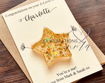 May include: A gold glitter star with the text "JULY CREATIONS" on it. The star is on a card with the text "Congratulations on your A-Level results Charlotte You're a star! Love from Mark & Sarah xx"