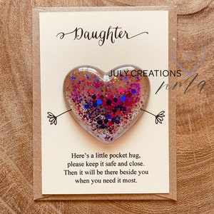 May include: A card with the word "Daughter" written in cursive on it. A small heart-shaped resin ornament with pink, blue, and red glitter is attached to the card. The card reads "Here's a little pocket hug, please keep it safe and close. Then it will be there beside you when you need it most."