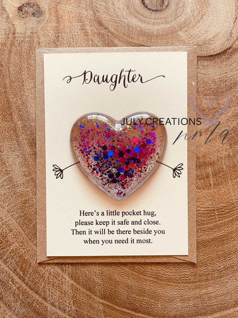 May include: A card with the word "Daughter" written in cursive on it. A small heart-shaped resin ornament with pink, blue, and red glitter is attached to the card. The card reads "Here's a little pocket hug, please keep it safe and close. Then it will be there beside you when you need it most."