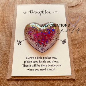 May include: A small heart-shaped resin ornament filled with iridescent glitter. The ornament is attached to a card with the text "Daughter" and "Here's a little pocket hug, please keep it safe and close. Then it will be there beside you when you need it most."