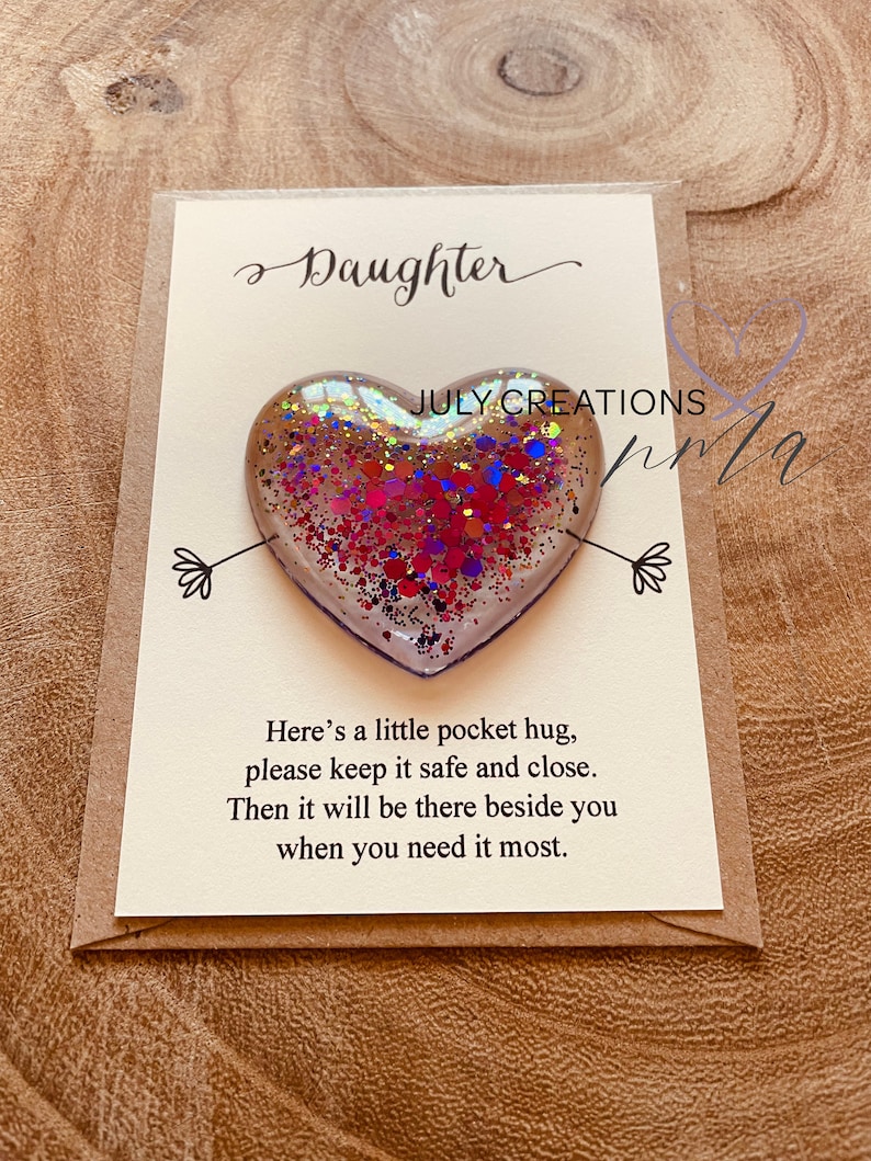 May include: A small heart-shaped resin ornament filled with iridescent glitter. The ornament is attached to a card with the text "Daughter" and "Here's a little pocket hug, please keep it safe and close. Then it will be there beside you when you need it most."