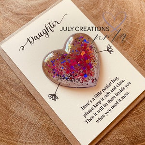 May include: A card with the text "Daughter" and "JULY CREATIONS" on it. A heart-shaped resin ornament with glitter and iridescent colors is attached to the card. The text on the card reads "Here's a little pocket hug, please keep it safe and close. Then it will be there beside you when you need it most."