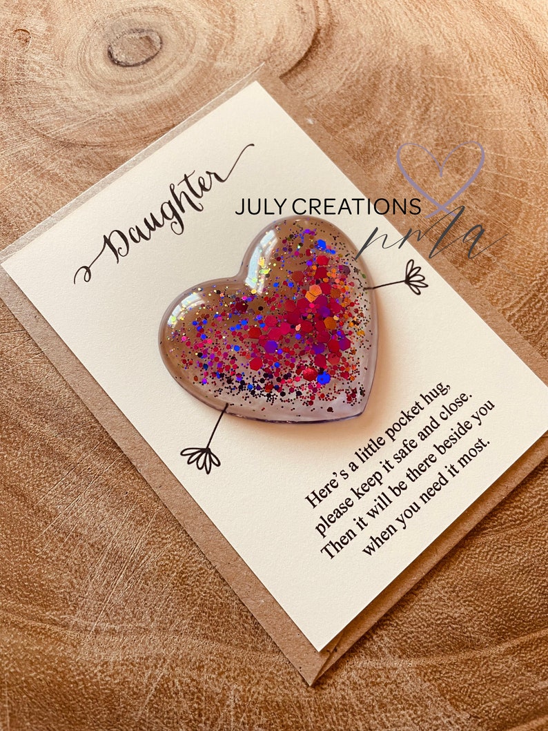 May include: A white card with the text "Daughter" and "JULY CREATIONS" in cursive. A clear heart-shaped resin ornament filled with colorful glitter is attached to the card with a small arrow. The card reads "Here's a little pocket hug, please keep it safe and close. Then it will be there beside you when you need it most."
