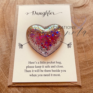 May include: A small heart-shaped resin ornament filled with red, pink, and blue glitter. The ornament is attached to a card with the text "Daughter" and "Here's a little pocket hug, please keep it safe and close. Then it will be there beside you when you need it most."