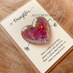 May include: A white card with the text "Daughter" and "JULY CREATIONS" in cursive. A clear heart-shaped resin ornament filled with colorful glitter is attached to the card with a small arrow. The card reads "Here's a little pocket hug, please keep it safe and close. Then it will be there beside you when you need it most."