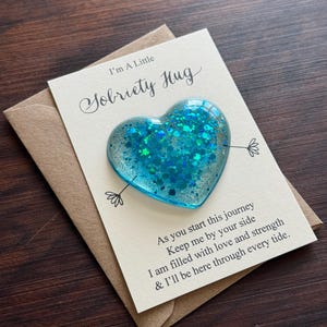 Può includere: Un ciondolo a forma di cuore turchese in resina con glitter, appoggiato su un biglietto e una busta. Il biglietto recita "I'm A Little Sobriety Hug" con un messaggio incoraggiante. Il ciondolo è largo circa 5 cm.
