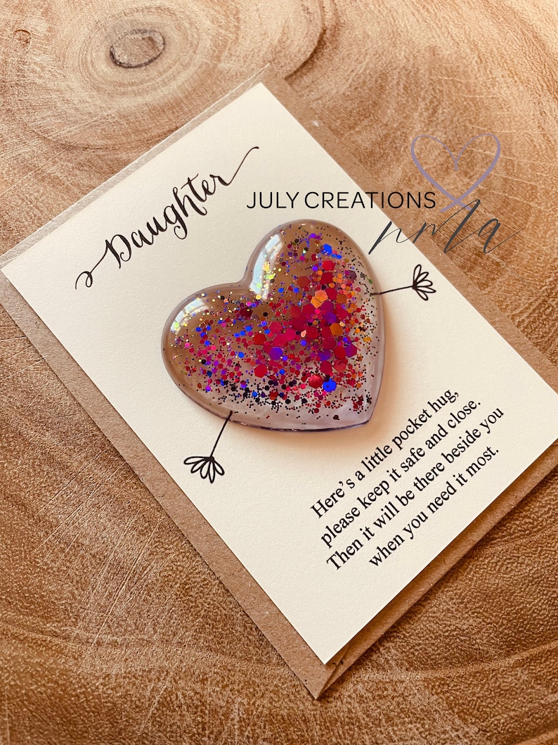 May include: A white card with the text "Daughter" and "JULY CREATIONS" in black cursive writing. A clear heart-shaped resin ornament filled with colorful glitter is attached to the card with a black arrow. The card reads "Here's a little pocket hug, please keep it safe and close. Then it will be there beside you when you need it most."