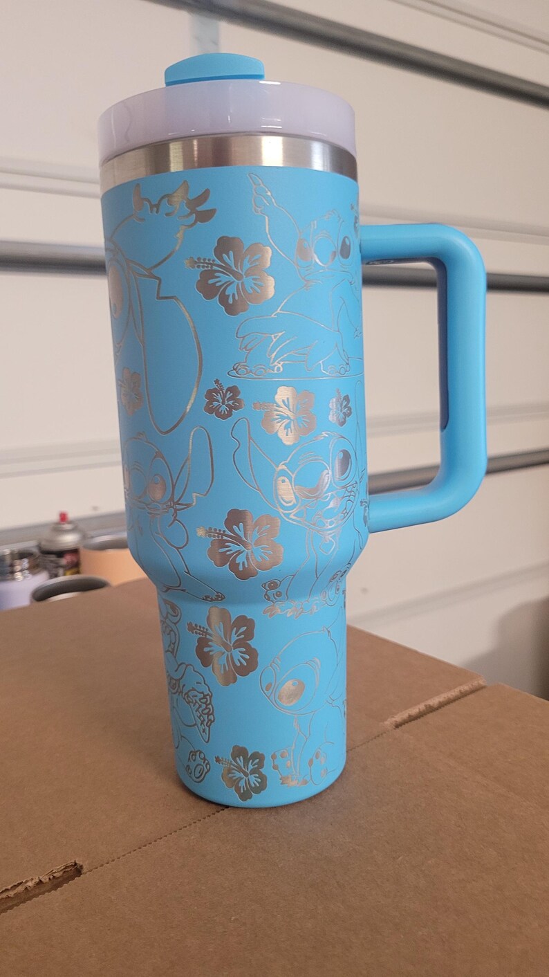 Puede incluir: Una taza de viaje de acero inoxidable azul claro con asa. La taza tiene un dise&ntilde;o plateado de Stitch de Lilo & Stitch y flores de hibisco.