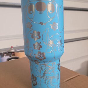 Puede incluir: Un vaso de acero inoxidable azul claro con una tapa blanca. El vaso tiene un dise&ntilde;o grabado en plata de Stitch y flores.