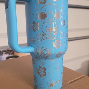 Puede incluir: Una taza de viaje de acero inoxidable azul claro con asa y tapa azules. La taza tiene un dise&ntilde;o plateado de Stitch de Lilo & Stitch y flores de hibisco.
