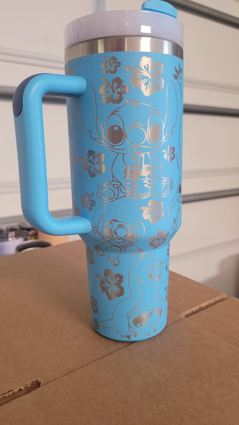 Puede incluir: Una taza de viaje de acero inoxidable azul claro con asa y tapa azules. La taza tiene un dise&ntilde;o plateado de Stitch de Lilo & Stitch y flores de hibisco.