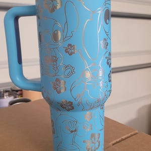 Puede incluir: Una taza de viaje de acero inoxidable azul claro con un asa azul y una tapa. La taza tiene un dise&ntilde;o plateado de Stitch de Lilo & Stitch y flores tropicales.