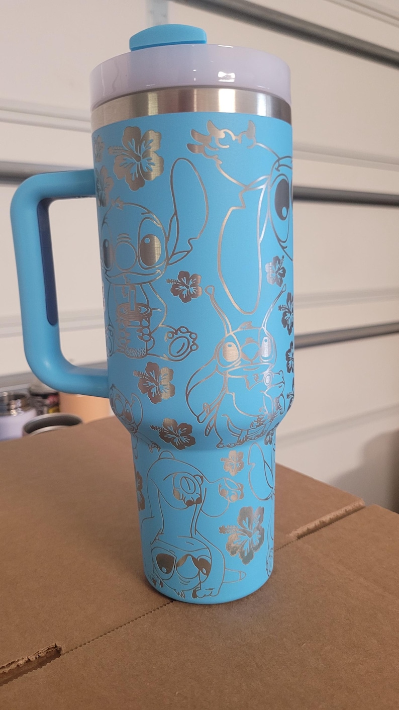 Puede incluir: Una taza de viaje de acero inoxidable azul claro con un asa azul y una tapa. La taza tiene un dise&ntilde;o plateado de Stitch de Lilo & Stitch y flores tropicales.