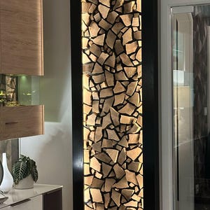 May include: A decorative wall panel featuring a mosaic of irregularly shaped wooden pieces. The wood has a light brown color, and the panel is framed by black borders. The panel is illuminated from behind, creating a warm glow.