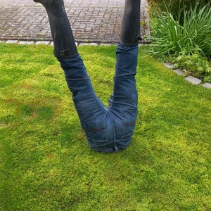 May include: A person's legs and torso are seen upside down, with the lower body buried in the grass. The person is wearing blue jeans and black rubber boots. The boots have a buckle detail. The scene is set on a green lawn.