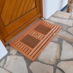 Puede incluir: Un felpudo de madera con un inserto de coco marrón. El felpudo está hecho de madera marrón claro y tiene un diseño de listones. El felpudo está colocado en un patio de piedra.