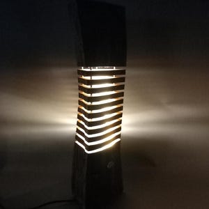 Puede incluir: Una lámpara de madera oscura con una base rectangular y una sección superior con listones iluminados. La lámpara emite un brillo cálido a través de las lamas horizontales. La lámpara está enchufada a una toma de corriente.