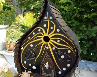 Puede incluir: Una casita para pájaros negra y dorada con un diseño de conchas y cangrejo. La casita tiene una pequeña puerta y una abertura redonda para que los pájaros puedan entrar. La casita está decorada con conchas y un cangrejo.